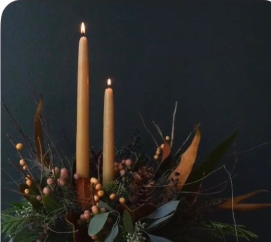 Evergreen arrangement with two amber taper candles lit against a dark wall
